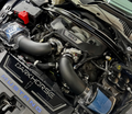 Close-up of a customized Ford Mustang engine bay featuring a large black intake pipe, a blue conical air filter, a clear-chambered HKS supercharger, and a black engine cover with 'DARKHORSE' and 'MUSTANG' branding.